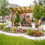 dark grey armor stone retaining wall with a flower bed built using the same material. A pergola has been built on an interlocking patio adjacent to the retaining wall.