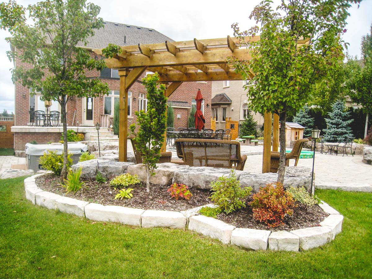 dark grey armor stone retaining wall with a flower bed built using the same material. A pergola has been built on an interlocking patio adjacent to the retaining wall.